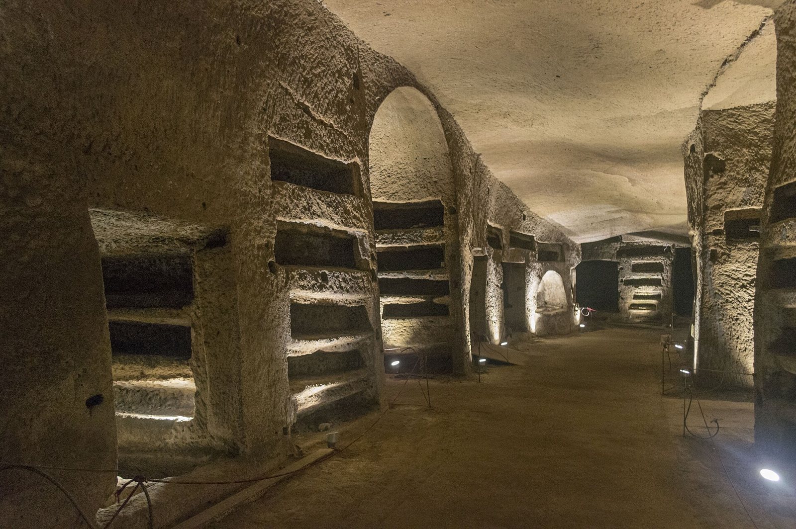 Galleria scavata nel tufo delle Catacombe di San Gennaro a Napoli, con file di loculi e arcate illuminate da luci basse