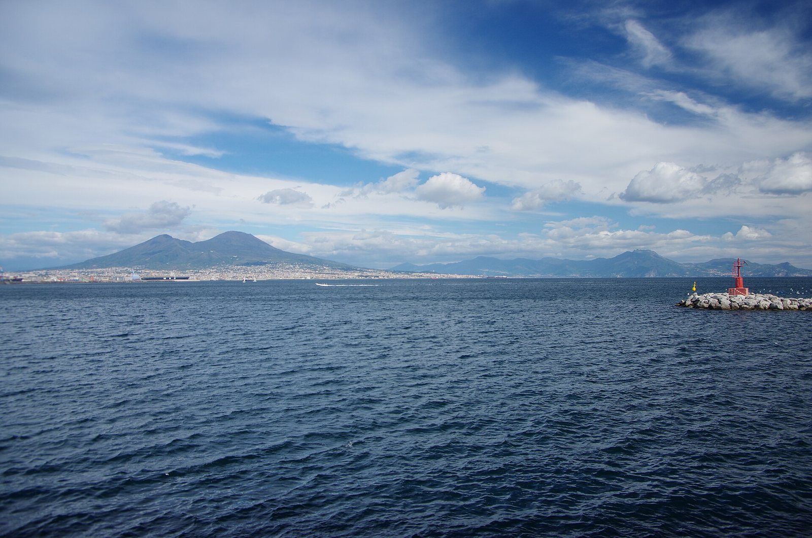 Il Golfo di Napoli visto dal mare con il Vesuvio sullo sfondo, la città lungo la costa e un piccolo faro rosso sulla scogliera a destra