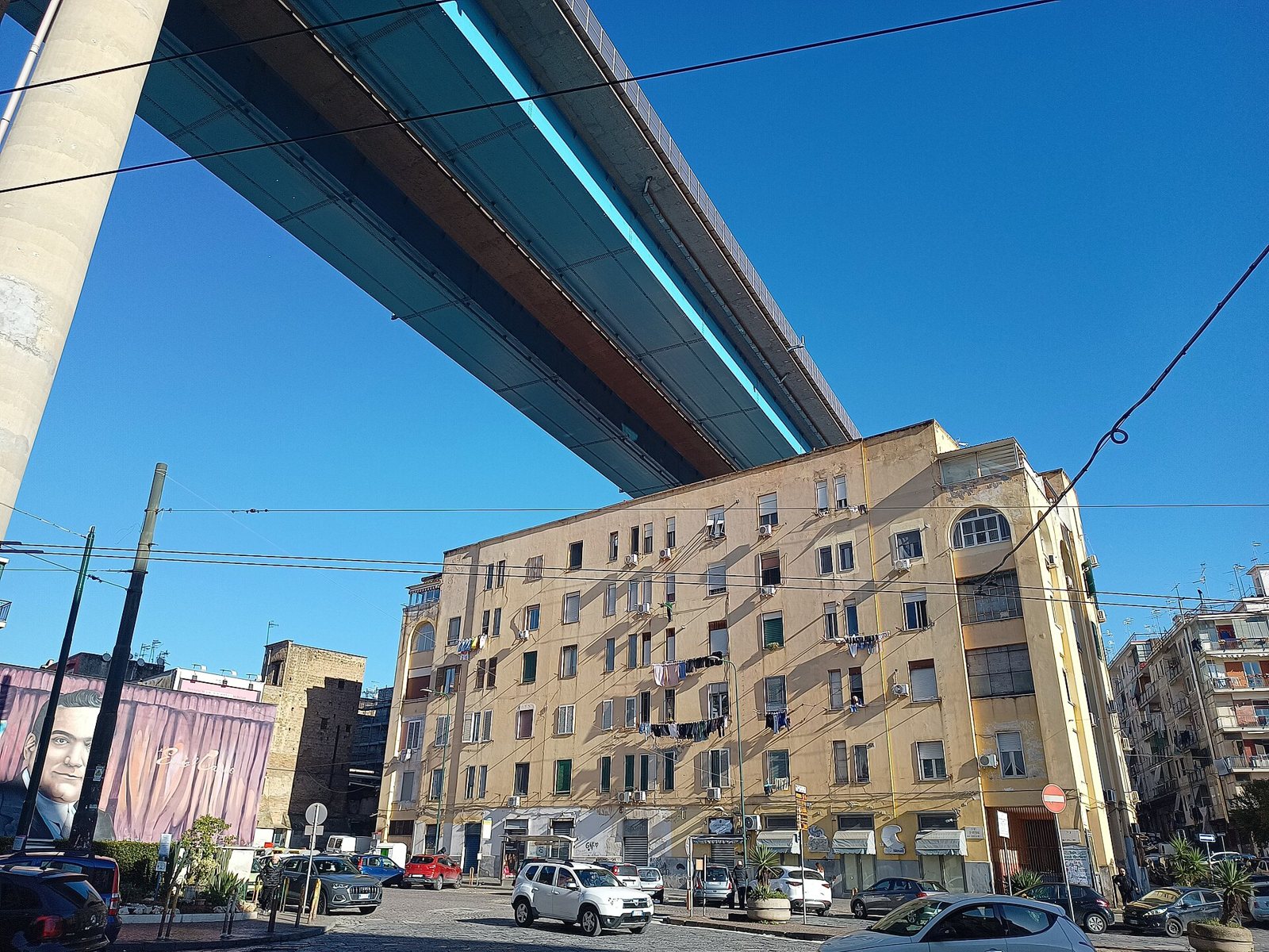 Viadotto sopraelevato che passa sopra piazza Ottocalli a Napoli, con un palazzo e auto in sosta sotto un cielo azzurro