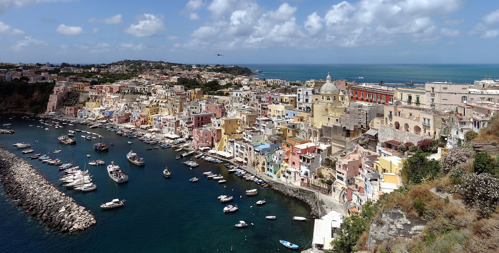 Veduta dall'alto della Marina Corricella a Procida, con le case colorate affacciate sul porticciolo pieno di barche e il mare sullo sfondo
