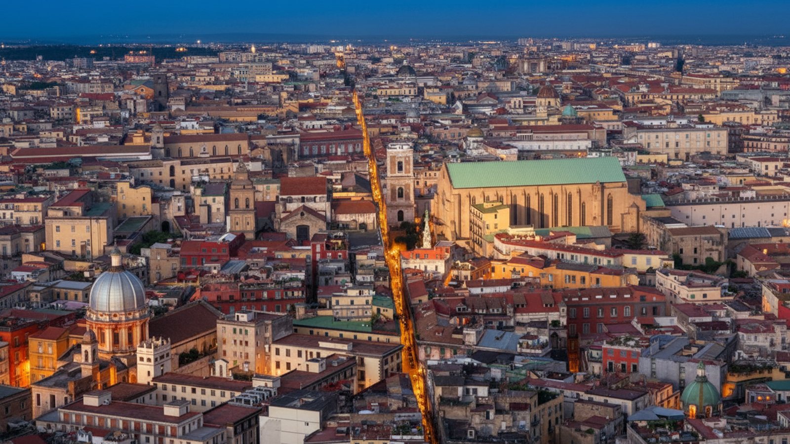 Veduta dall'alto del centro storico di Napoli al crepuscolo, tagliato in linea retta dalla strada illuminata di Spaccanapoli tra tetti, cupole e campanili